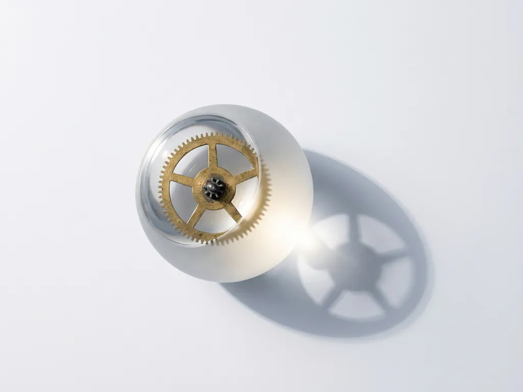 Messing tandwiel verbonden met doorschijnende glazen bol met zacht licht, geometrische schaduwen op wit oppervlak