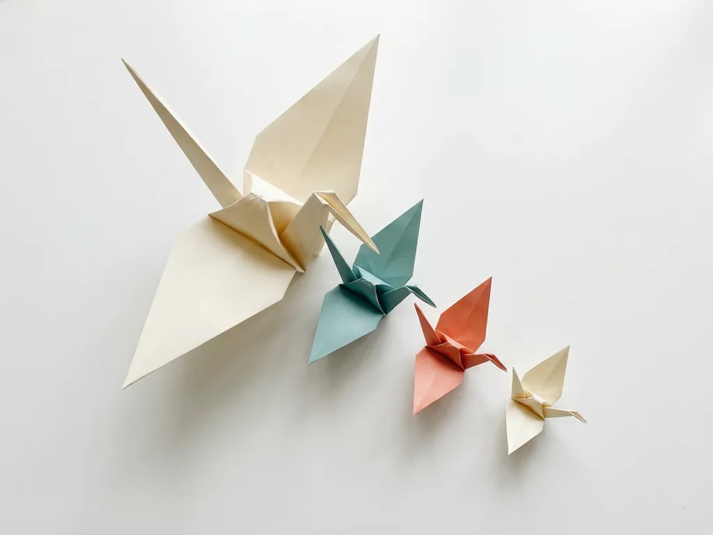 Origami kraanvogel die ontvouwt in drie kleinere kraanvogels op een diagonale lijn, zachte schaduwen op wit oppervlak, blauwgroen en koraal accenten.