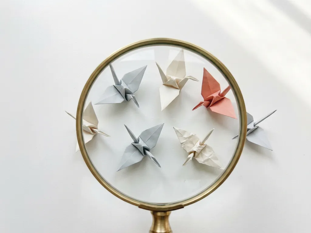 Vergrootglas boven papieren origami vogels in grijs en ivoor tinten met koraal accent, sommige perfect gevouwen, andere met subtiele imperfecties.